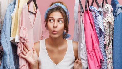 A woman with a playful expression stands between racks of colorful clothes against a pink background. She wears a blue headband and a white top.