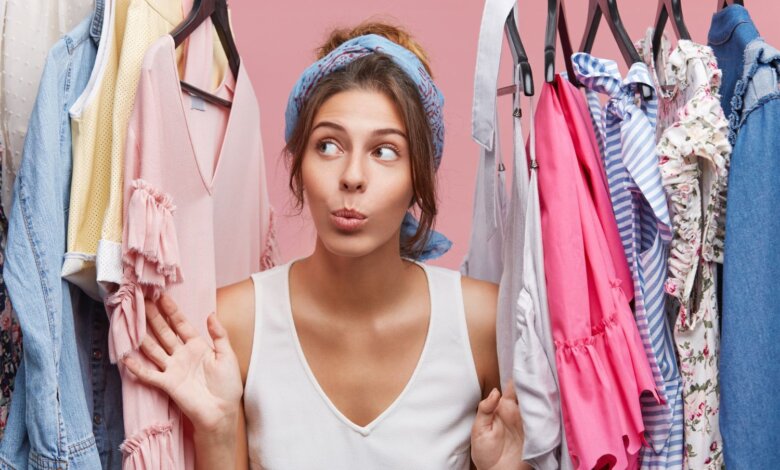 A woman with a playful expression stands between racks of colorful clothes against a pink background. She wears a blue headband and a white top.