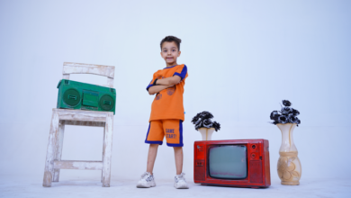 A young child in an orange outfit with arms crossed stands in front of a white chair with a green boombox, a red vintage TV, and two vases with black flowers.