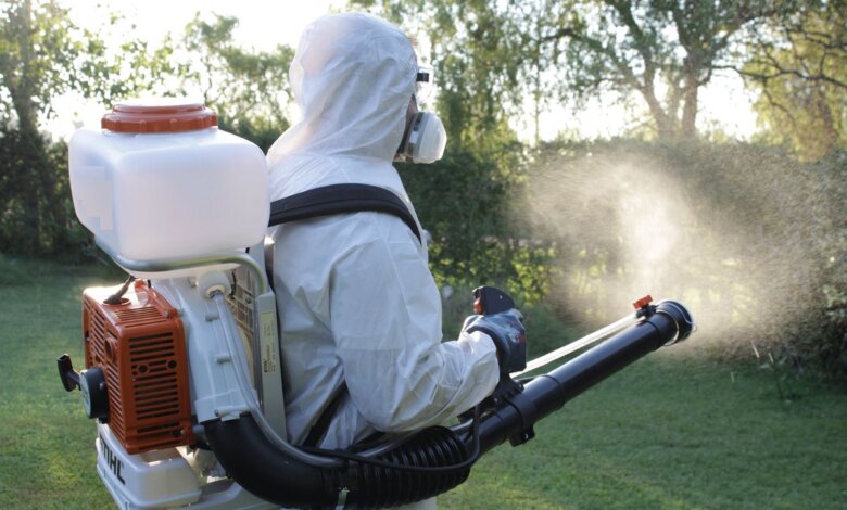 A pest control technician in a full protective suit sprays chemicals outdoors using a professional mist blower in a garden or yard.