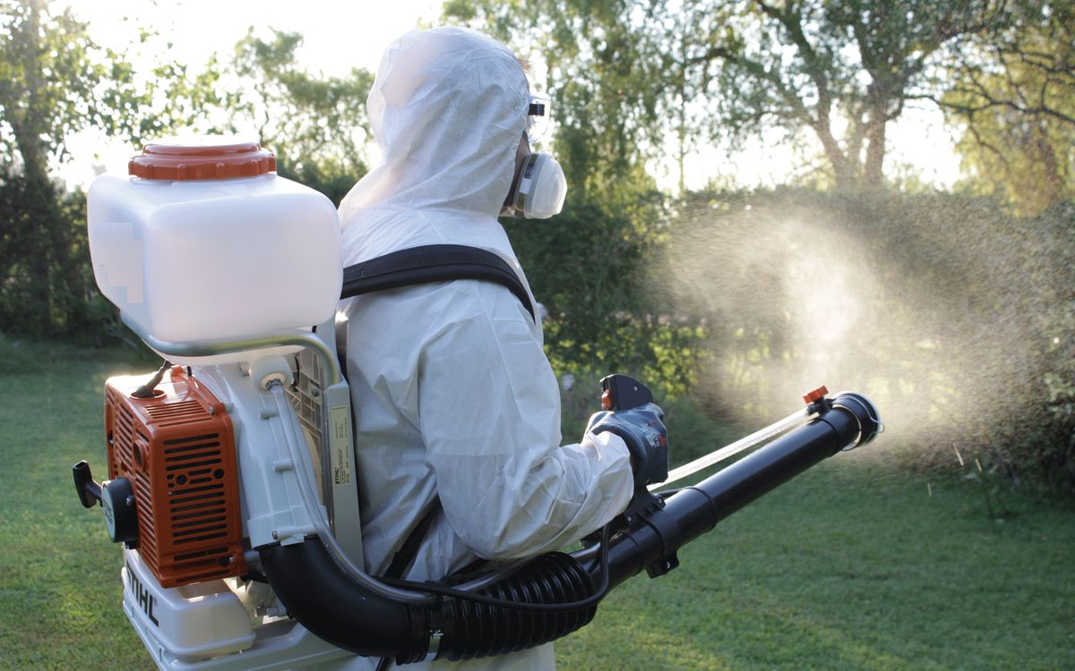 A pest control technician in a full protective suit sprays chemicals outdoors using a professional mist blower in a garden or yard.