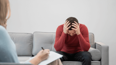 A man sits on a couch, resting his head in his hands, conveying a sense of stress or deep contemplation.