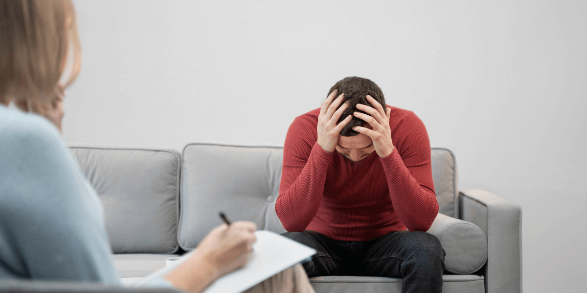 A man sits on a couch, resting his head in his hands, conveying a sense of stress or deep contemplation.